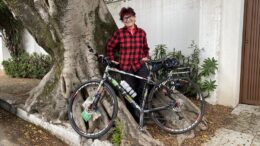 renata falzoni com camisa xadrez vermelha e preta, enconstada em uma grande árvore, sorri para a foto com sua bicicleta.