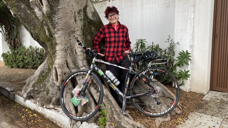 renata falzoni com camisa xadrez vermelha e preta, enconstada em uma grande árvore, sorri para a foto com sua bicicleta.