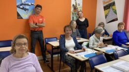 uma sala de aula com carteiras e 6 senhoras sentadas, em pé junto à parede do fundoo um homem oriental e uma jovem professora.,