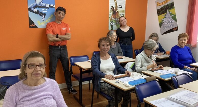 uma sala de aula com carteiras e 6 senhoras sentadas, em pé junto à parede do fundoo um homem oriental e uma jovem professora.,