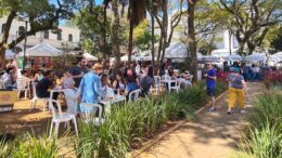 foto de um evento anterior, com mesinhas de plástico na praça e muitas pessoas, além de barraquinhas ao fundo