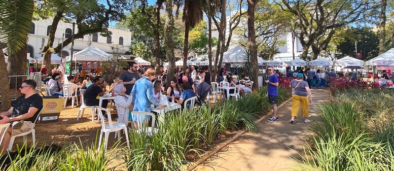 foto de um evento anterior, com mesinhas de plástico na praça e muitas pessoas, além de barraquinhas ao fundo