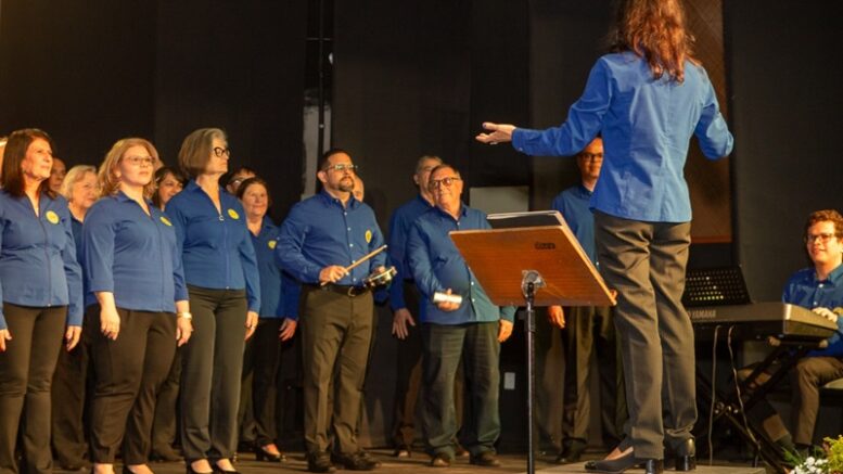 imagem de um coral com os componentes em pé de calça marrom e blusa azul, a regente, de costas com a mesma roupa e no canto se vê um sorridente tecladista.