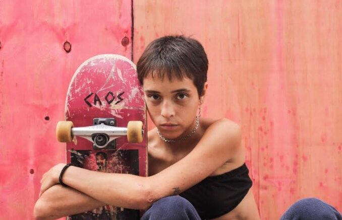 imagem de skatista menina, de cabelos curtos,abraçando seu skate encostada em uma parede rosa.