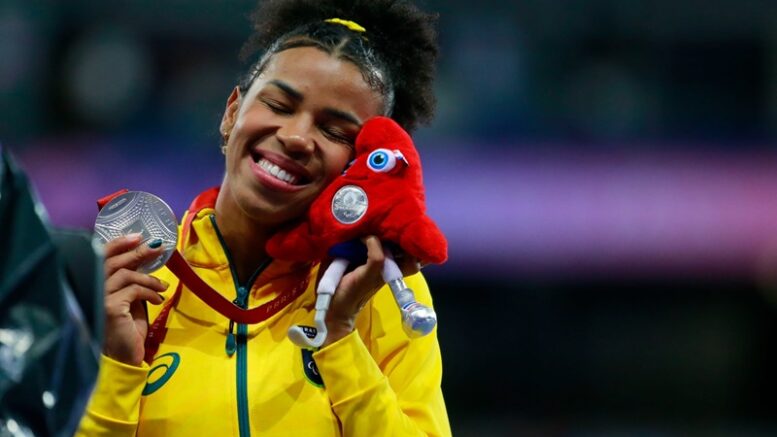 Rayane Soares, atleta brasileira em close no pódio, de olhos fechados e um largo sorriso, mostrando a medalha de prata e com o rosto colado na pelúcia do mascote paralimpico.