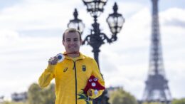 atleta mostra a medalha e a pelúcia dos jogos, sobre uma ponte antiga com a torre eifel ao fundo.