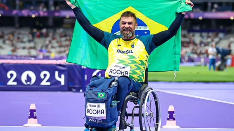 atleta andre rocha, cadeirante, sorri com a bandeira do Brasil erguida com os braços atrás de si.