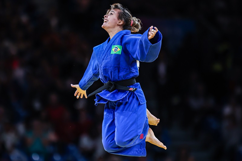 atleta de judo com quimono azul salta de alegria com os braços abertos e as pernas flexionadas