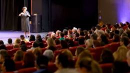 em um teatro com a plateia cheia, visto do fundo para o palco, uma mulher sobre ele fala ao microfone;