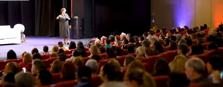 em um teatro com a plateia cheia, visto do fundo para o palco, uma mulher sobre ele fala ao microfone;