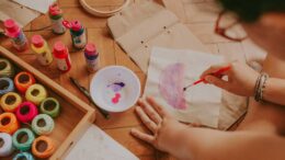 Imagem vista de cima de uma pessoa pintando com tinta aquarelável, ao lado uma caixa baixa de madeira com mutos novelinhos que poderam ser