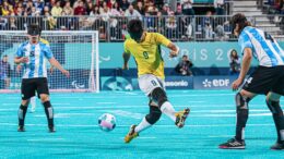 imagem de jogo de futebol de cegos, com dois argentinos um de cada lado e ao centro um vbrasileiro chutando a bola