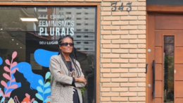 Djamila Ribeiro, mulher negra de cabelo longo trançado, posa em frente ao espaço feminismos plurais, uma casa de tijolinhos aparente de uma grande janela de vidro com o nome ravado no vidro.