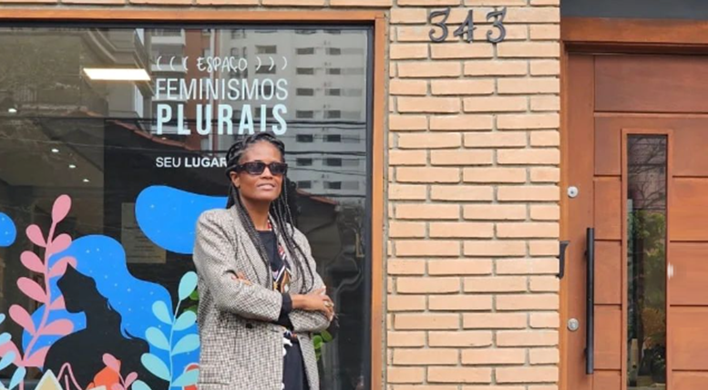 Djamila Ribeiro, mulher negra de cabelo longo trançado, posa em frente ao espaço feminismos plurais, uma casa de tijolinhos aparente de uma grande janela de vidro com o nome ravado no vidro.