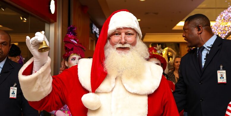 imagem de papai noel com um sininho, em pé em um corredor com dois seguranças ao seu lado e uma elfa logo atrás