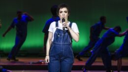 atriz interpretando elis, de macacão jeans e camiseta branca, atrás dela, no palco escurecido e com luz verde, se veem alguns bailarinos em jeans também.