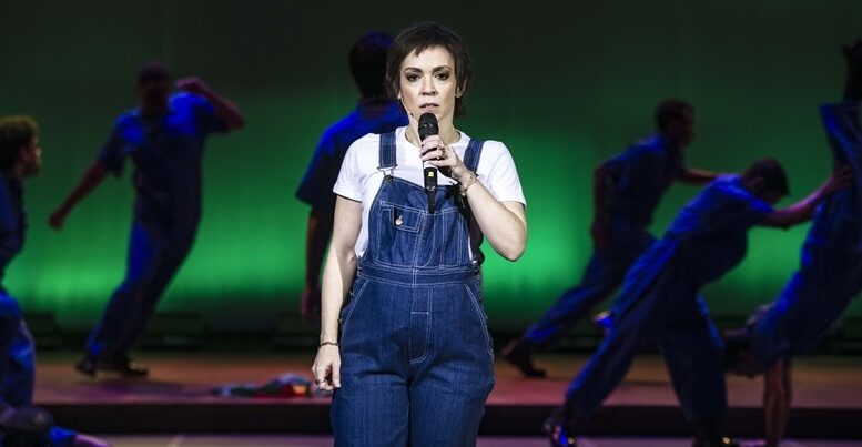 atriz interpretando elis, de macacão jeans e camiseta branca, atrás dela, no palco escurecido e com luz verde, se veem alguns bailarinos em jeans também.