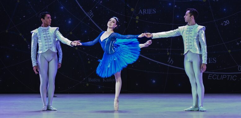 bailarina com vestido de tutu azul, fica na ponta, com uma das pernas esticada atrás, e dá as mãos a dois bailarinos, ao lado dela.