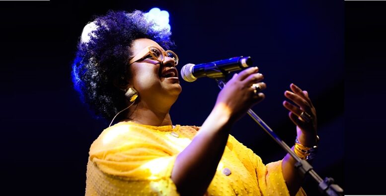 imagem da cantora de lado, cantando, segurando o microfone em um pedestal. ela está com uma blusa amarela, é negra e usa óculos.