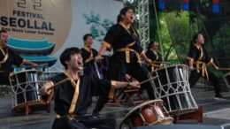 grupo de percussão japonesa em um palco. seis componentes vestindo kimonos escuros, gritando com olhos fechados e braços abertos em frete a seus tambores.