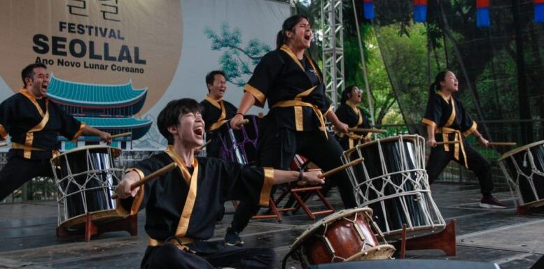 grupo de percussão japonesa em um palco. seis componentes vestindo kimonos escuros, gritando com olhos fechados e braços abertos em frete a seus tambores.