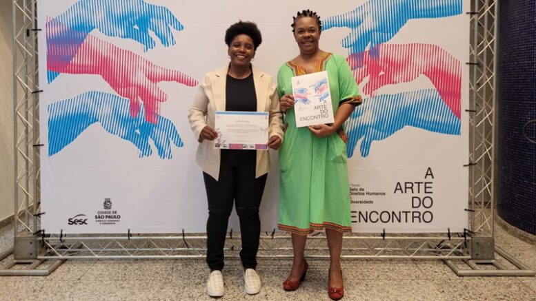 imagem de duas representantes da ONG mostrando os premios em frente a um backdrop do programa da prefeitura