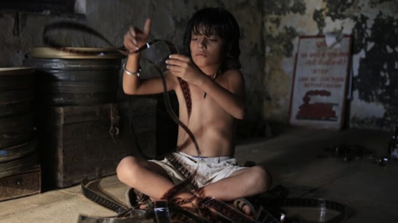 imagem de um menino sem camisa, sentado no chão de uma sala pouco iluminada, de pernas cruzadas vendo negatovos de filmes