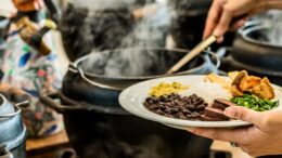 imagem fechada de mãos femininas se servindo de feijoada em caldeirão fumegante.