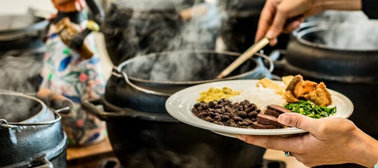 imagem fechada de mãos femininas se servindo de feijoada em caldeirão fumegante.