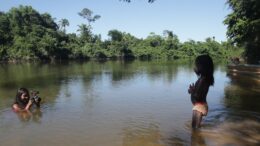 em um rio, uma indígena com uma câmera, abaixada na água, filma uma criança.