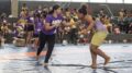 duas atletas de wrestling lutando em pé em uma área de jogo, com plateia sentada atrás