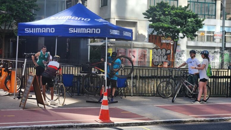 tenda azul montada na praça do ciclista,, com uma bike erguida no equipamento e dois ciclistas aguardando sua vez.
