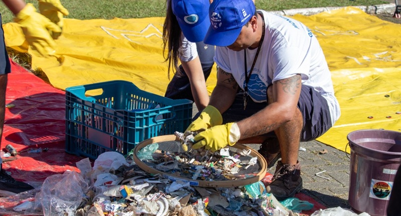 dois adultos de boné azul e camiseta branca, agaixados separam lixo plastico depositado sobre uma lona amarela, sob o sol