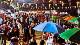 imagem noturna de festa junina, oom bandeirinhas e muitas pessoas em pé em um piso de terra. e luzinhas ao fundo.