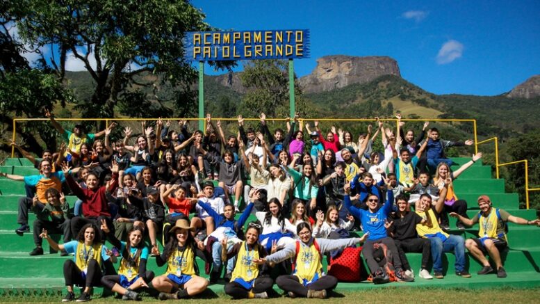 arquibancada ao ar livre com vários jovens e ao fundo a pedra do baú, uma planca indica acampamento paiol grande