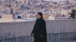 Imagem de mulher vestida de preto, num fim de tarde ao lado de um muro com a cidade vista atrás em um morro