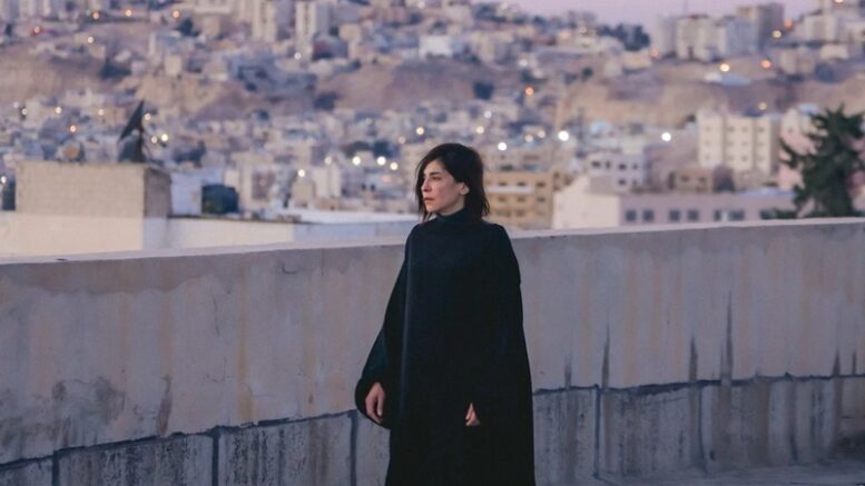 Imagem de mulher vestida de preto, num fim de tarde ao lado de um muro com a cidade vista atrás em um morro