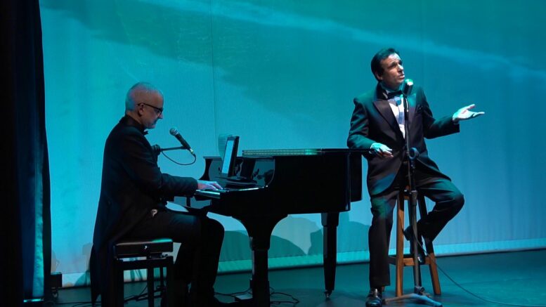 imagem com luz azul, um pianista de fraque tocando um piano de calda e um cantor em um banco alto, com o cotovelo apoiado no piano.