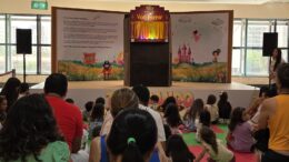 imagem de um palco de teatro montando num final de corredor com grandes janelas atras e um pouco de publico de costas para a foto, em frente a ele.