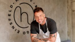 chef e autor do livro, debruçado sobre uma mesa. ele veste avental e na parede está a logo do seu restaurante.