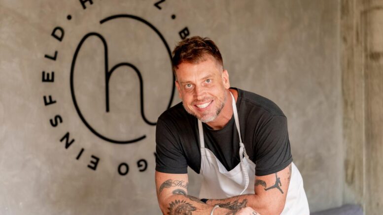 chef e autor do livro, debruçado sobre uma mesa. ele veste avental e na parede está a logo do seu restaurante.