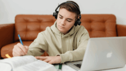 menino sentado no chão, em frente a uma mesa baixa com notebook e cadernos. ele usa fones de ouviro e está escrevendo à lápis em um dos cadernos.