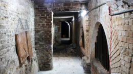 imagem de passagem subterrânea com paredes de tijolo e portas em arco.