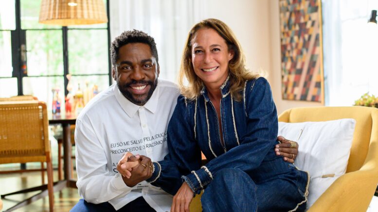 Lázaro Ramos e Andrea Beltrão sentados em uma sala, posando de mãos dadas e sorrindo para a câmera.