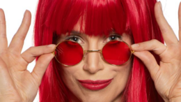 Mel Lisboa em close com figurino de Rita Lee, segurando óculos rodondos..