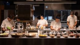 imagem horizontal de uma cozinha com quatro chefes em duas linhas de bancadas e fogões