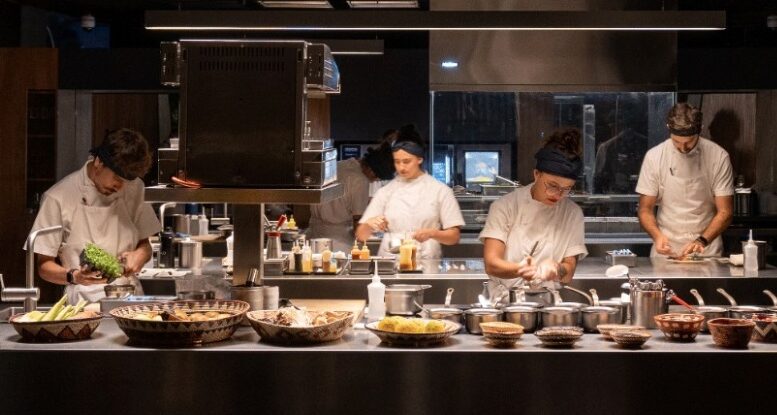 imagem horizontal de uma cozinha com quatro chefes em duas linhas de bancadas e fogões