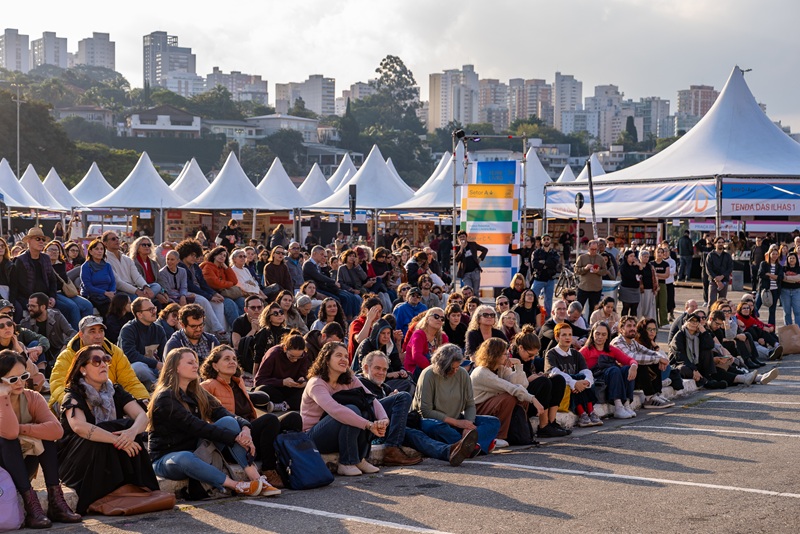 imagem diurna em que se vê muitas pessoas sentdas no chão, atrás delas várias tendas brancas e mais ao fundo árvores e prédios vistos ao longe