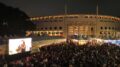 imagem noturna da Feira do Livro na praça Charles Muller com o estádio ao fundo e muitas pessoas sentadas no chão no gramado.