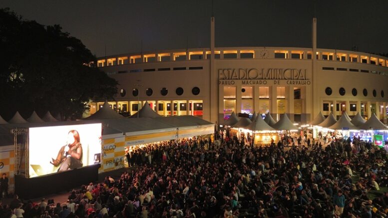 imagem noturna da Feira do Livro na praça Charles Muller com o estádio ao fundo e muitas pessoas sentadas no chão no gramado.